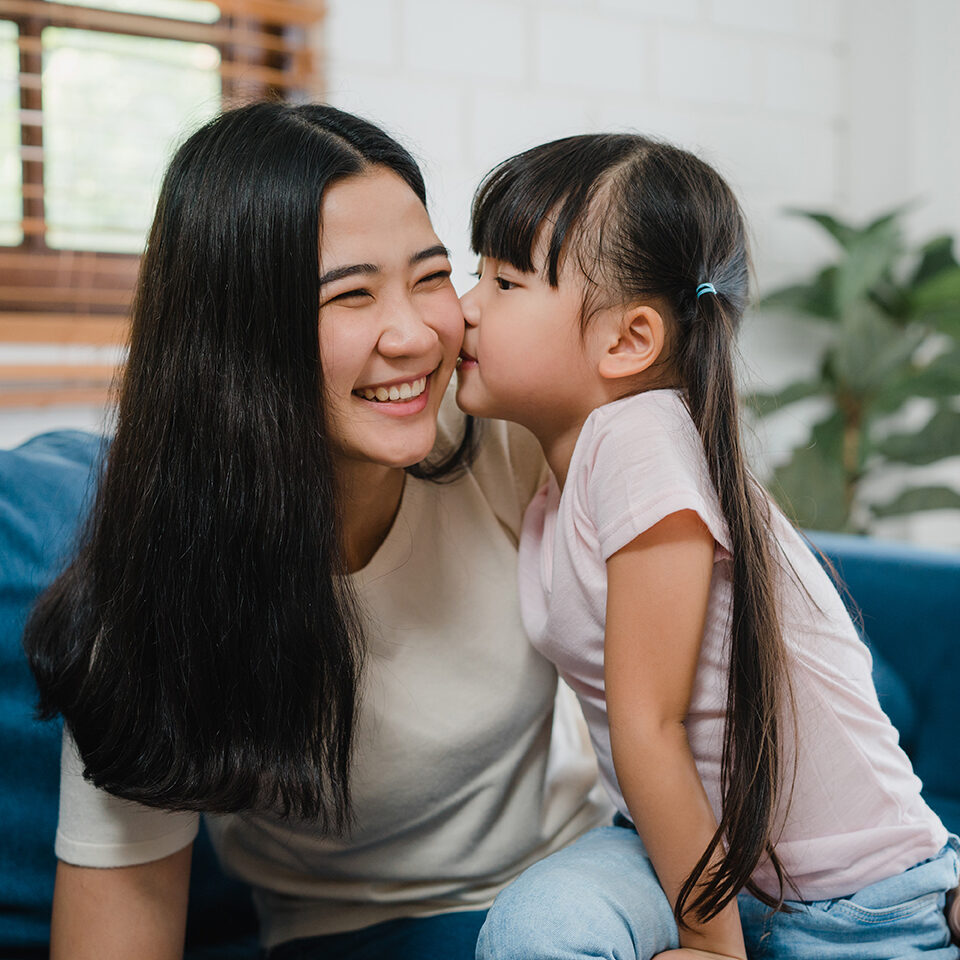Happy Asian family mom and daughter embracing kissing on cheek congratulating with birthday at house. Self-isolation, stay at home, social distancing, quarantine for coronavirus prevention.